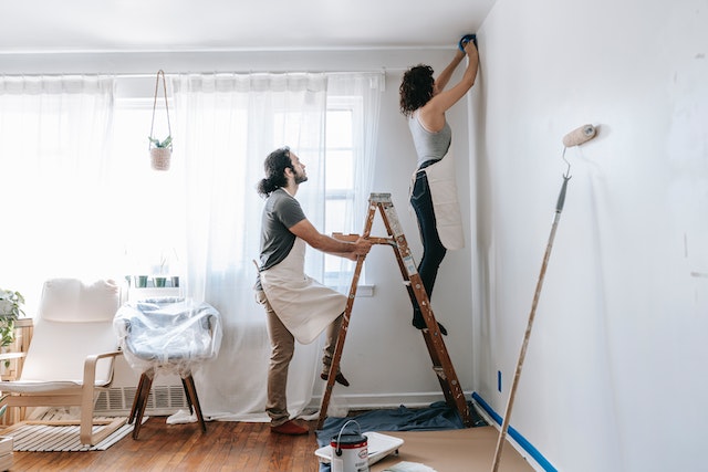 a contractor with long dark hair and brown pants holds a ladder while another contractor with curly brown hair and a white apron applies painters tape to a wall